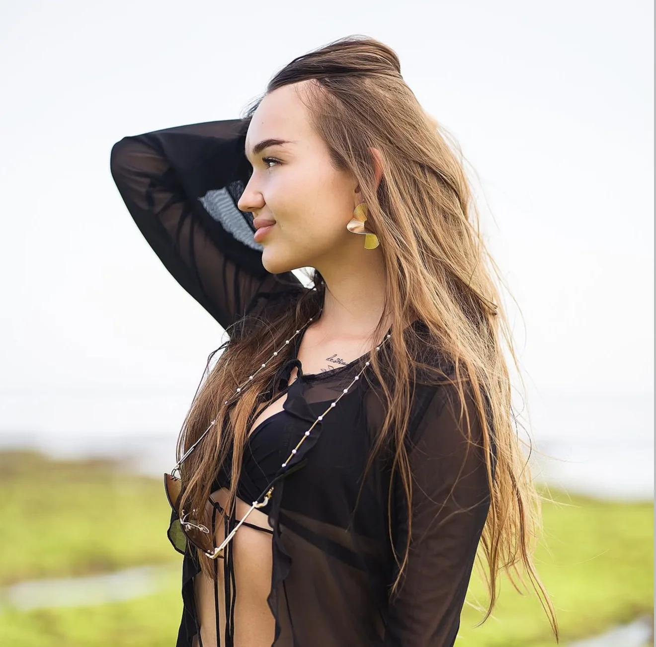 Woman with long hair in a sheer black shirt, one arm raised behind her head, looking away into bright daylight.